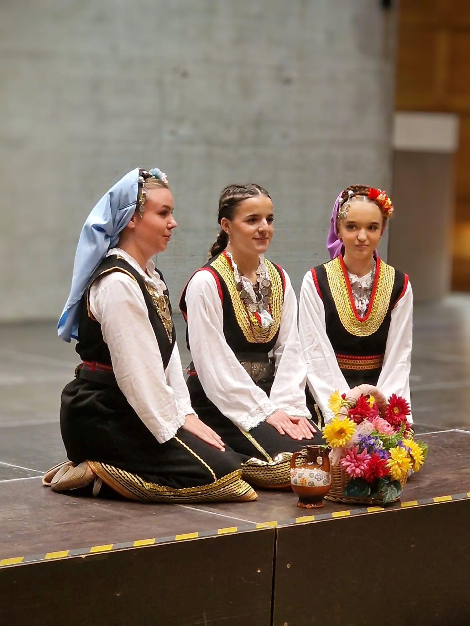 Drei Tänzerinnen in serbischer Tracht knien auf der Bühne neben einem Blumenkorb